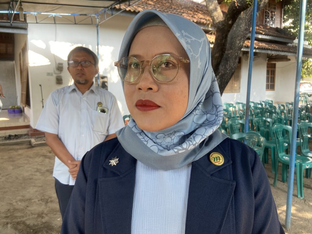 Close-up of a woman wearing a blue floral hijab and glasses, with a man in a light shirt and badge in the background at an outdoor event.