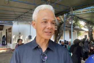 Close-up of a smiling man with white hair and glasses hanging from his shirt, at an outdoor gathering under a blue framework canopy.