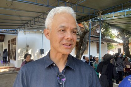 Close-up of a smiling man with white hair and glasses hanging from his shirt, at an outdoor gathering under a blue framework canopy.
