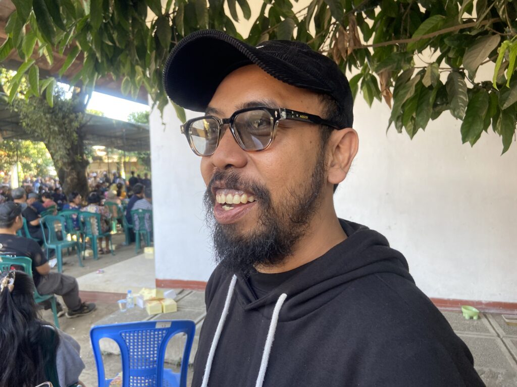 Close-up portrait of a man wearing a black cap and glasses, smiling, with an outdoor gathering and green plastic chairs in the background.