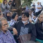 Crowd of elderly people seated outdoors at a shaded gathering, many with masks around their necks, some looking at papers or phones beneath a tree backdrop.