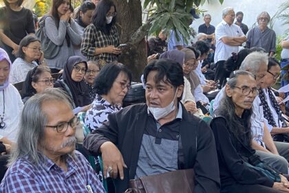 Crowd of elderly people seated outdoors at a shaded gathering, many with masks around their necks, some looking at papers or phones beneath a tree backdrop.