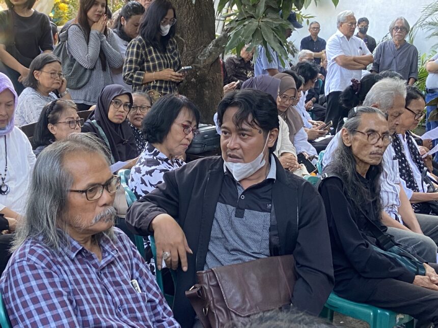 Crowd of elderly people seated outdoors at a shaded gathering, many with masks around their necks, some looking at papers or phones beneath a tree backdrop.