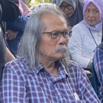 Older man with gray hair and glasses wearing a plaid shirt sits among a crowd of people, some in headscarves, outdoors.