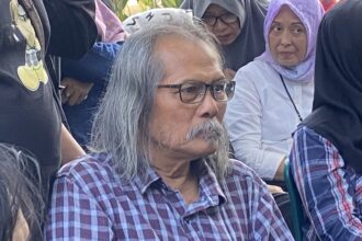 Older man with gray hair and glasses wearing a plaid shirt sits among a crowd of people, some in headscarves, outdoors.