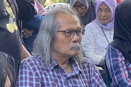 Older man with gray hair and glasses wearing a plaid shirt sits among a crowd of people, some in headscarves, outdoors.