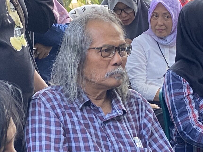 Older man with gray hair and glasses wearing a plaid shirt sits among a crowd of people, some in headscarves, outdoors.