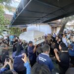 Large crowd surrounding a ceremonial coffin being carried through an outdoor space under a blue canopy, with many attendees taking photos or watching solemnly.