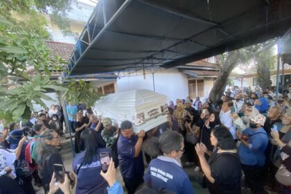 Large crowd surrounding a ceremonial coffin being carried through an outdoor space under a blue canopy, with many attendees taking photos or watching solemnly.