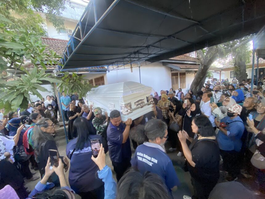 Large crowd surrounding a ceremonial coffin being carried through an outdoor space under a blue canopy, with many attendees taking photos or watching solemnly.