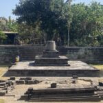 Stone pedestal monument on a tiered platform in a sunny park, with two people sitting on the steps and dense greenery behind the scene.