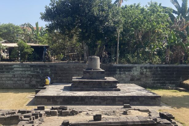 Stone pedestal monument on a tiered platform in a sunny park, with two people sitting on the steps and dense greenery behind the scene.
