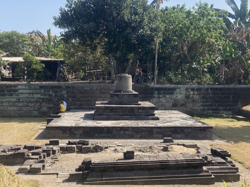 Stone pedestal monument on a tiered platform in a sunny park, with two people sitting on the steps and dense greenery behind the scene.