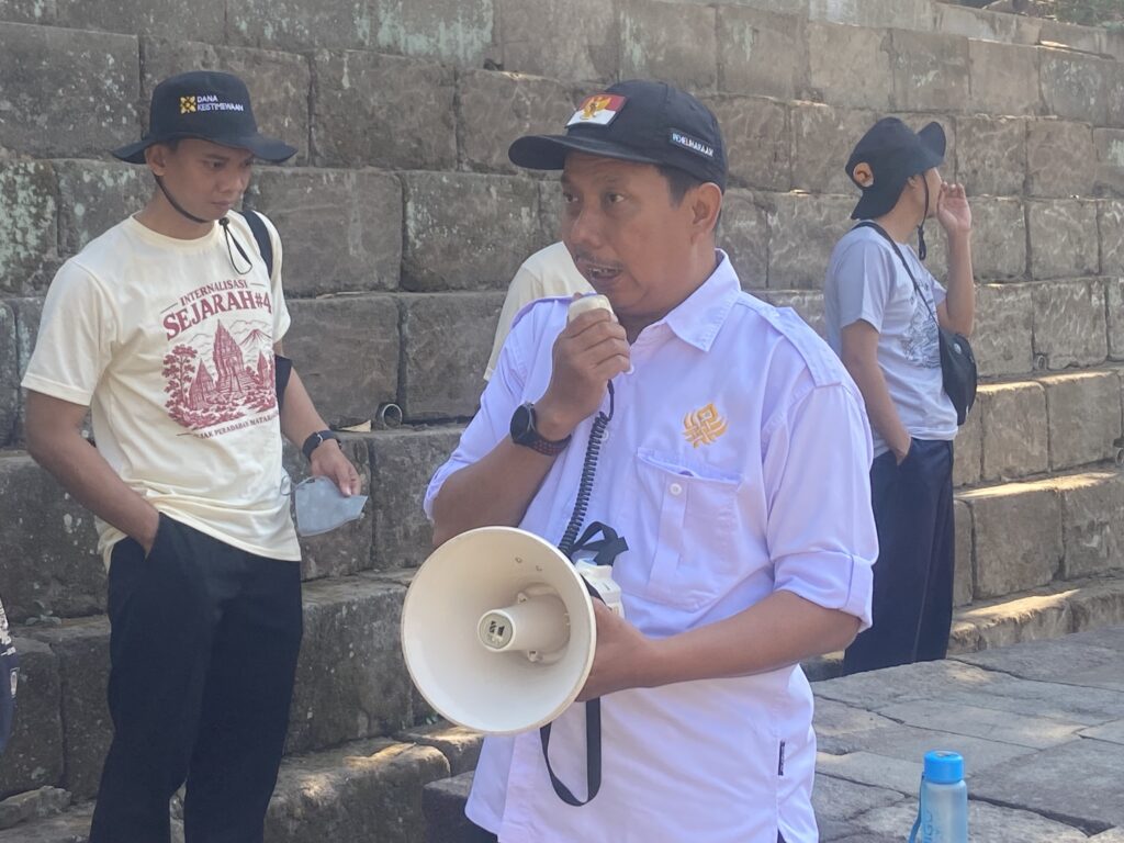 IMG_4239 - Mabur.co Man in light shirt holding a megaphone outdoors near stone steps; others stand nearby with hats.