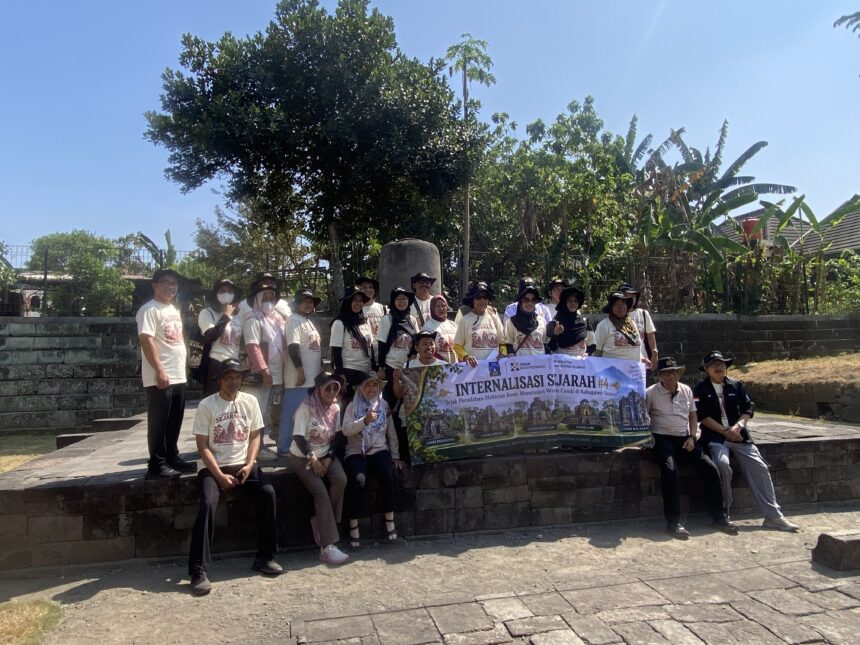 Group of people in matching light-colored shirts posing on stone steps outdoors, holding a large banner for a group event under a sunny sky.
