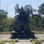 Stone tiered monument in a park, surrounded by trees and a clear blue sky behind it.