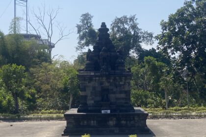 Stone tiered monument in a park, surrounded by trees and a clear blue sky behind it.