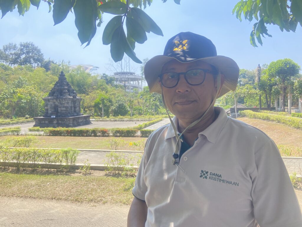 Man wearing a wide-brimmed hat and glasses posing in a sunny garden with a stone shrine in the background, logo visible on his shirt.