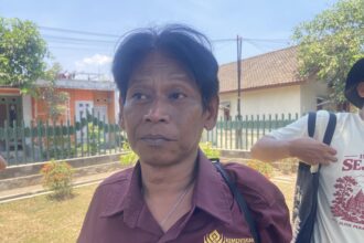Close-up portrait of a woman in a maroon shirt outdoors, with a green fence and houses in the background.