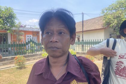 Close-up portrait of a woman in a maroon shirt outdoors, with a green fence and houses in the background.
