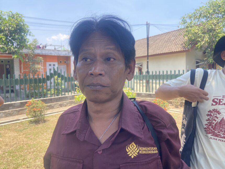 Close-up portrait of a woman in a maroon shirt outdoors, with a green fence and houses in the background.