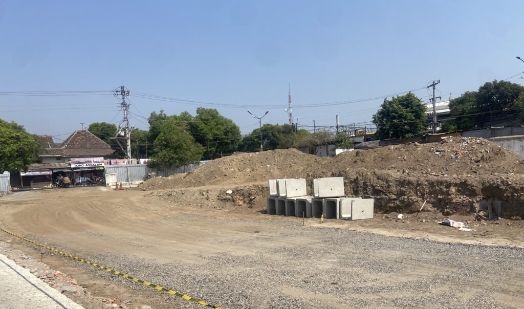 Construction site with dirt piles, stacked concrete blocks, and a dirt road next to a small shop named Toko Assalam under a clear blue sky.