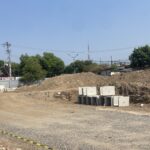 Construction site with dirt piles, stacked concrete blocks, and a dirt road next to a small shop named Toko Assalam under a clear blue sky.