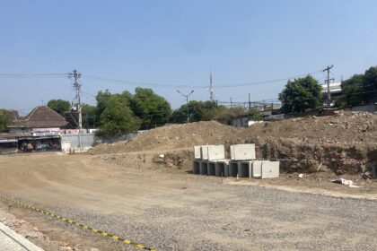 Construction site with dirt piles, stacked concrete blocks, and a dirt road next to a small shop named Toko Assalam under a clear blue sky.
