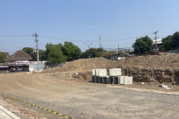 Construction site with dirt piles, stacked concrete blocks, and a dirt road next to a small shop named Toko Assalam under a clear blue sky.