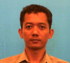 Portrait of a man with short dark hair, wearing a light brown shirt, facing the camera against a blue backdrop.