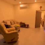 Cozy living room with beige sofa and matching recliner, small coffee table, and cross hanging above a doorway on a warm-lit wall