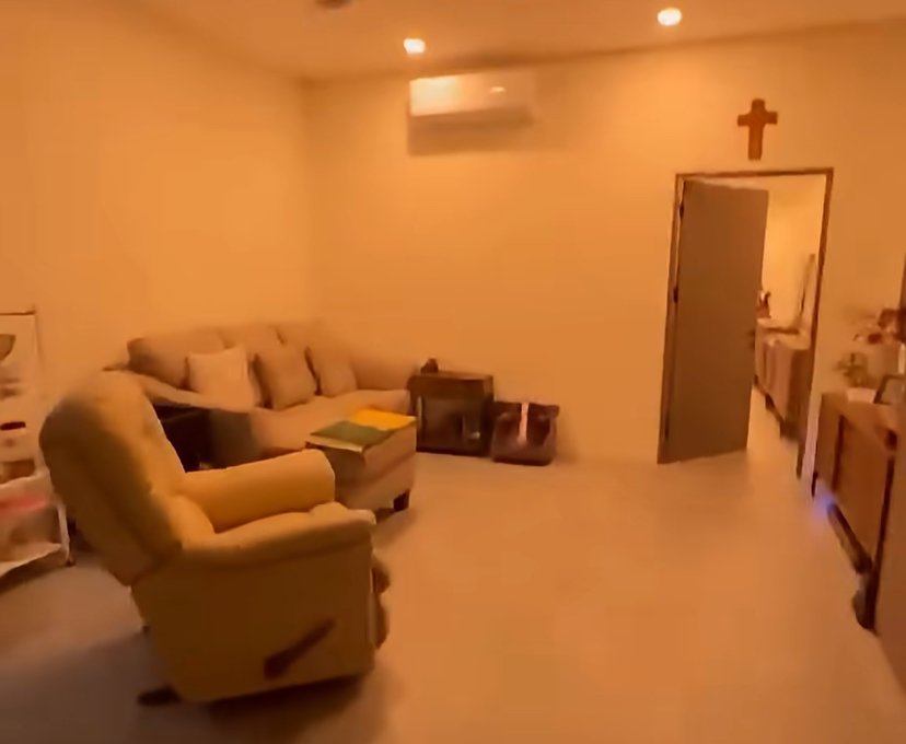 Cozy living room with beige sofa and matching recliner, small coffee table, and cross hanging above a doorway on a warm-lit wall