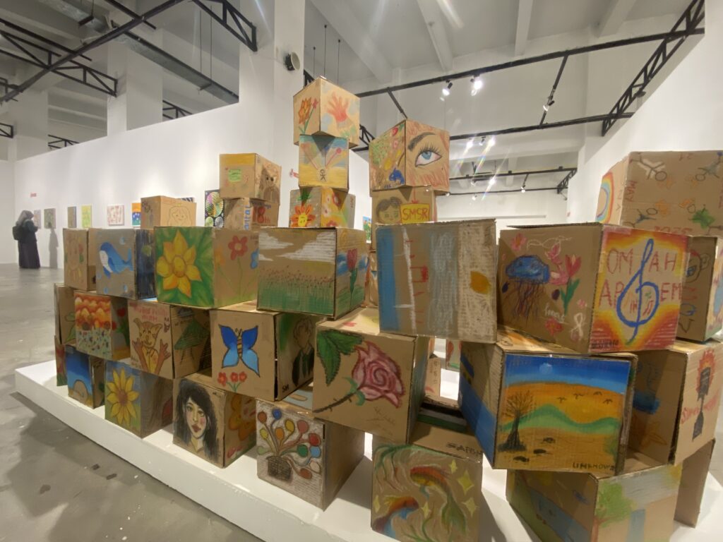 IMG_4379 - Mabur.co Colorful painted cardboard boxes stacked in a sculptural tower inside a white-walled gallery, with a person viewing artworks in the background.