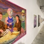 Large colorful painting of people in striped clothing hanging on a white gallery wall, with a pink frame; other artworks line the corridor.