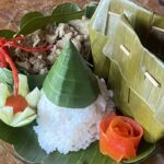 Banana leaf pocket opened to reveal sticky rice with savory filling, garnished with cucumber and carrot rose on a wooden plate.