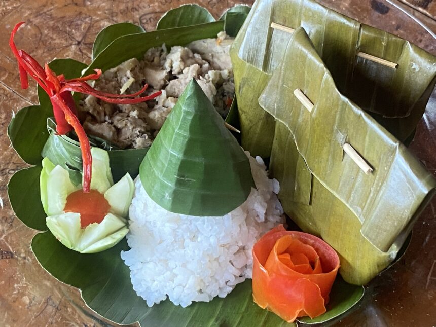 Banana leaf pocket opened to reveal sticky rice with savory filling, garnished with cucumber and carrot rose on a wooden plate.