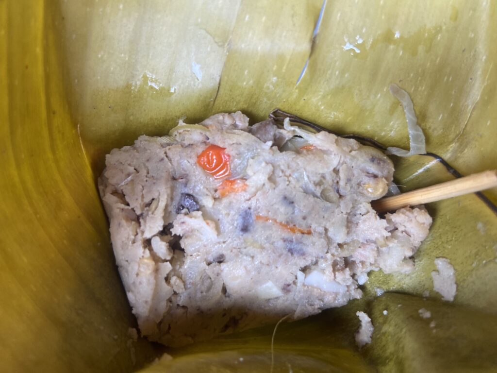 Close-up of a crumbly grayish dumpling with small orange bits inside a banana leaf wrapper.