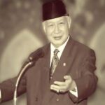 Elderly man in a suit and dark hat giving a public speech at a podium, gesturing with his right hand.