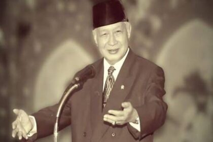 Elderly man in a suit and dark hat giving a public speech at a podium, gesturing with his right hand.