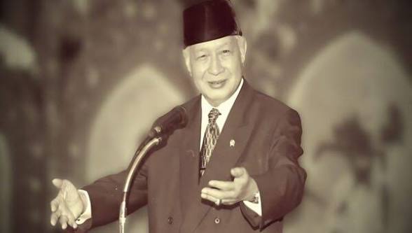 Elderly man in a suit and dark hat giving a public speech at a podium, gesturing with his right hand.