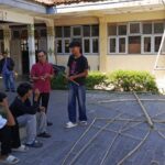 Group of students outside a school talking, with bamboo poles laid out on the pavement and a ladder in the background.
