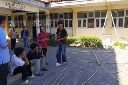Group of students outside a school talking, with bamboo poles laid out on the pavement and a ladder in the background.