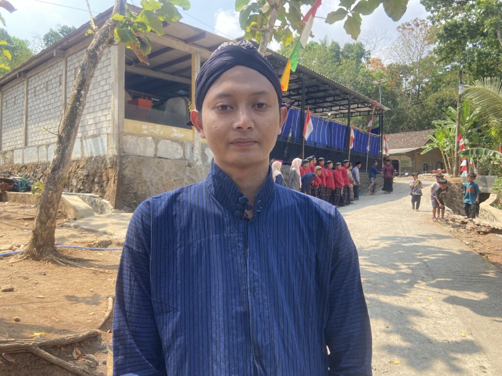 Self-portrait of a person in a blue striped shirt and dark headwrap, outdoors with a group of people and flags in the background.