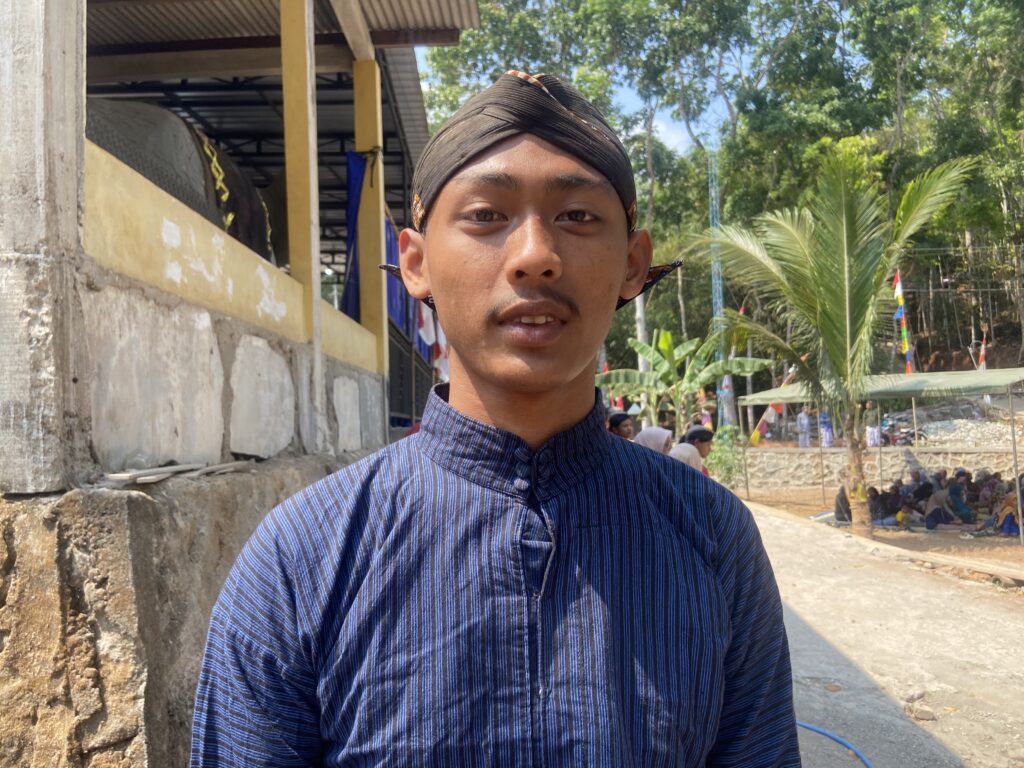 Young man taking a selfie outdoors in sunlight, wearing a blue striped shirt and a black headwrap; a yellow wall and trees behind.
