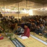 Large group of people in traditional clothing seated on mats under a canopy for a ceremony or gathering.