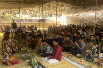 Large group of people in traditional clothing seated on mats under a canopy for a ceremony or gathering.