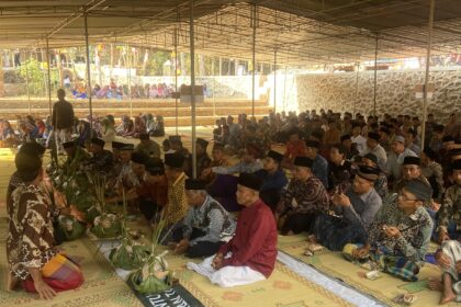Large group of people in traditional clothing seated on mats under a canopy for a ceremony or gathering.