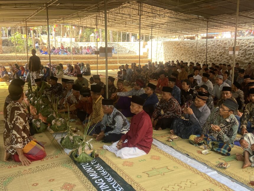 Large group of people in traditional clothing seated on mats under a canopy for a ceremony or gathering.