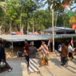 People in traditional clothing carrying woven baskets at an outdoor festival with colorful flags and stalls under trees.