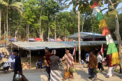 People in traditional clothing carrying woven baskets at an outdoor festival with colorful flags and stalls under trees.